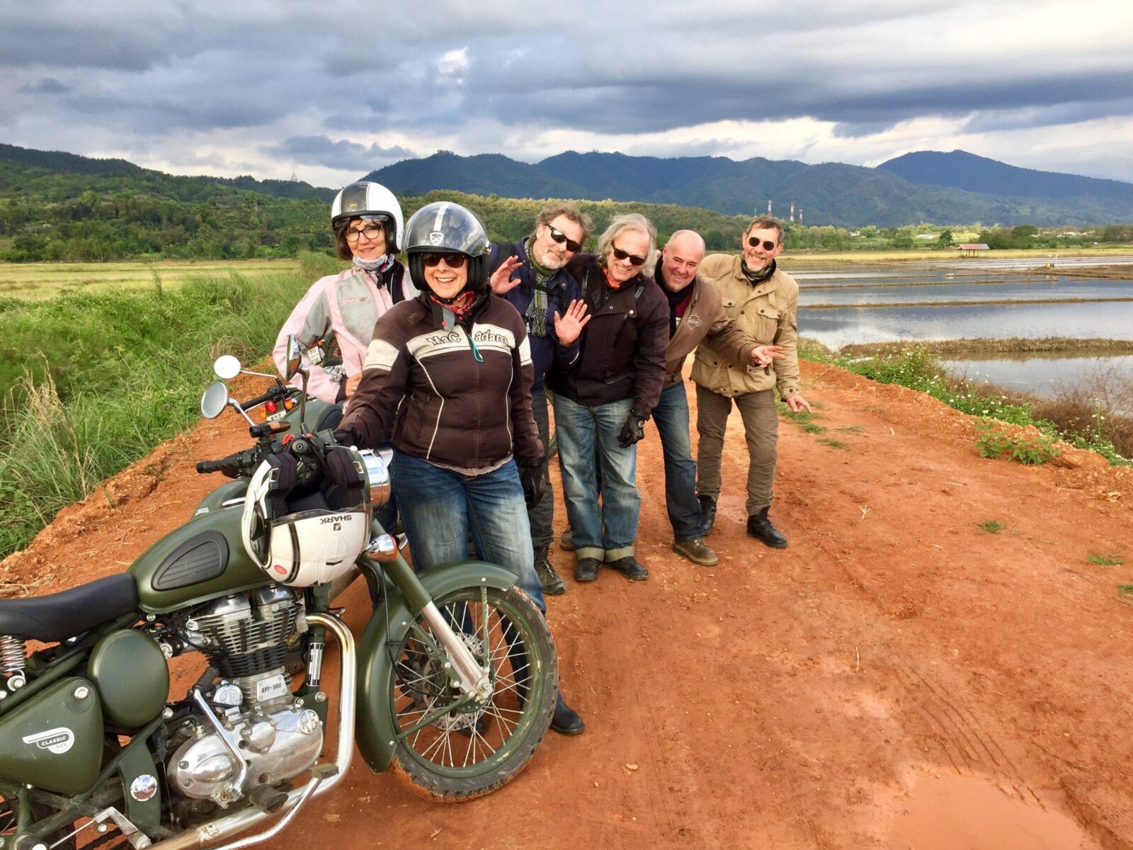 Group of smiling riders standing next to their Royal Enfield motorcycles beside a lake in northern Thailand
