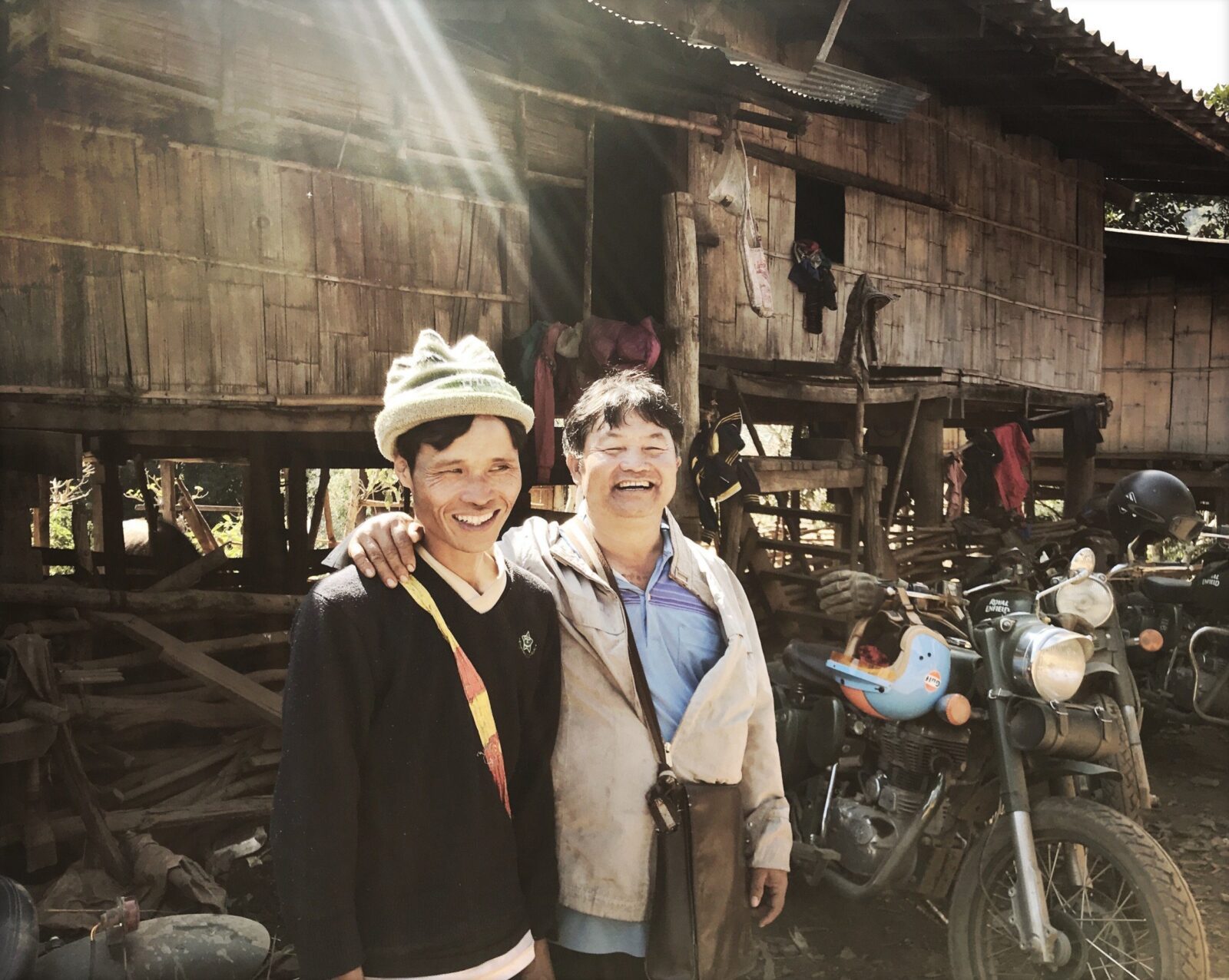 Two smiling villagers posing in front of a wooden house next to Royal Enfield motorcycles in northern Thailand